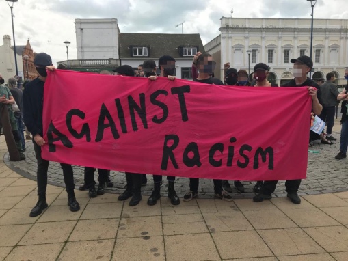 Image shows a banner saying 'Against Racism' held by a group of people dressed in black